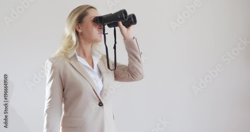 Modern beautiful young woman Caucasian in a business suit stands on a light background and looks through binoculars