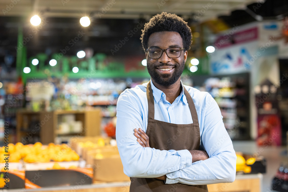 Fototapeta premium Young african american grocery store owner wearing an apron, arms crossed and smiling confidently at the camera while managing his small business among fresh produce