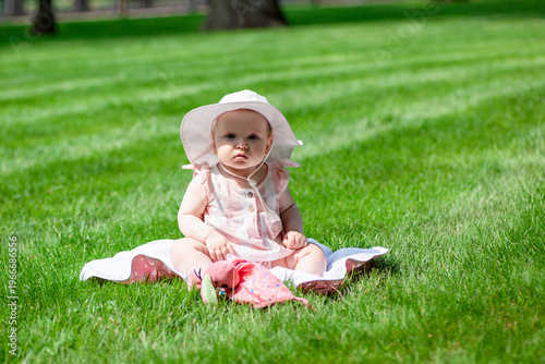 A child sits on a soft blanket in the grass. The child wears a sun hat and looks content. Sunlight shines down on the grassy area around.