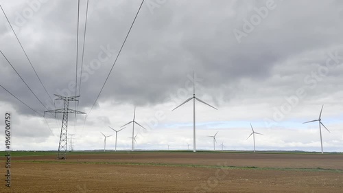 Expansive farmland scene with turbines rising above freshly tilled soil while a transmission line runs parallel to the field, linking production to the grid beneath a tense cloud filled sky