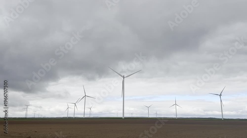 Wide view of brown plowed field with twelve active wind turbines under uncertain sky with clouds opening to blue, distant power line pylons visible in rural France