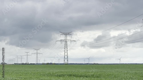 Perspective view where a row of transmission poles guides the eye across a green wheat field toward distant wind turbines, under a soft clouded sky shaping the rural energy landscape
