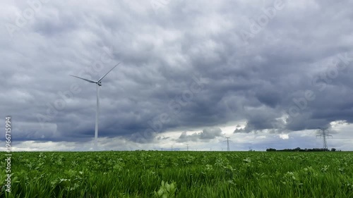 Lone turbine standing above open farmland beneath a dramatic sky before a storm, with distant transmission lines stretching across the horizon and structuring the rural landscape