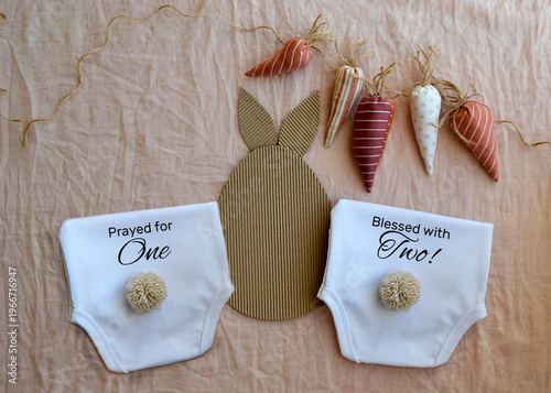 Two baby diapers with bunny tail and carrot garland for twin pregnancy announcement. Cute newborn clothing layout with Easter bunny theme reflecting double miracle birth celebration.