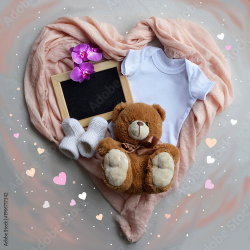 Baby announcement flat lay with chalkboard, white bodysuit, booties and teddy bear. Newborn outfit and toy inside heart shaped cloth frame. Pregnancy reveal concept for social media.