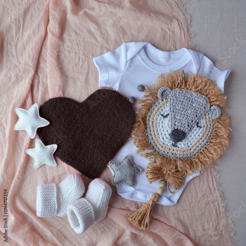 Baby bodysuit with crochet lion toy, heart and knitted booties on soft fabric. Newborn set for baby shower invitation. Cute infant clothing theme, nursery decoration and gifts.