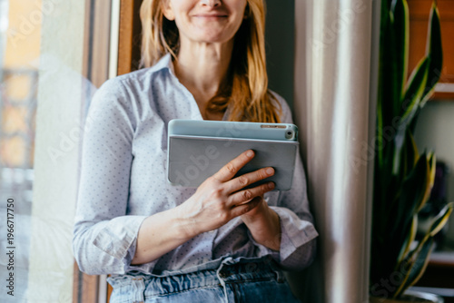 Foto Smiling caucasian woman in her 50s holding tablet near window in home interior