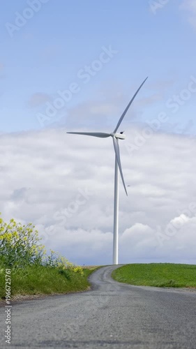 Portrait view of a wind turbine aligned at the end of a country path with green fields and a patch of yellow rapeseed, illustrating renewable energy in rural landscape in Vendée France