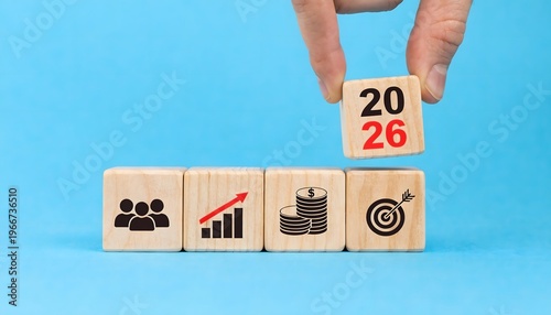 Hand placing a 2026 wooden block on a row of business icons including team, growth graph, money, and target on a light blue background. Concept of new year business goals and financial planning.