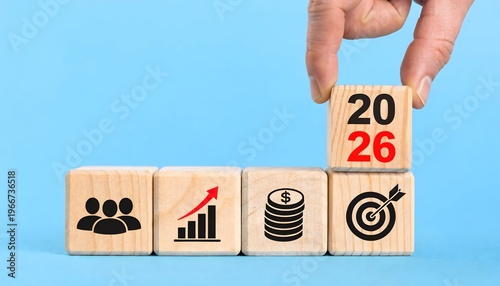 Hand placing a 2026 wooden block on a row of business icons including team, growth graph, money, and target on a light blue background. Concept of new year business goals and financial planning.