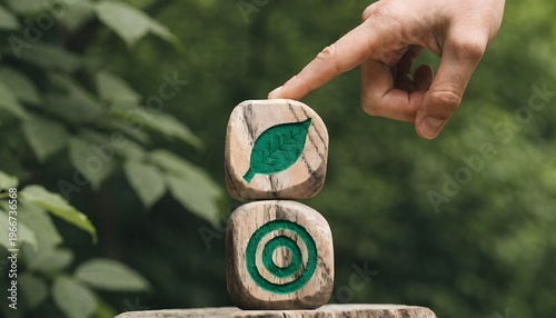 A hand with a finger touching a stack of two wooden blocks. Top block has a green leaf, bottom block has a green target symbol. Rustic wooden table, lush blurred green background. Concept of environme