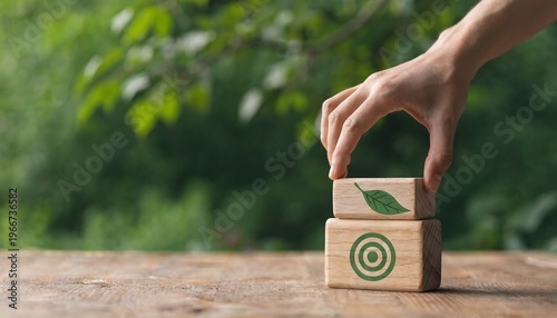 A hand with a finger touching a stack of two wooden blocks. Top block has a green leaf, bottom block has a green target symbol. Rustic wooden table, lush blurred green background. Concept of environme