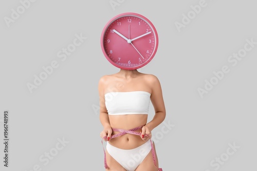 Young woman with clock instead of her head measuring waist on grey background...