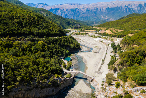 Aerial view of the historic Ottoman Katiu Bridge and circular turquoise thermal baths at the entrance of Langarica Canyon near Permet, Albania, set against the backdrop of the rugged Nemercka