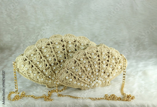 Beige crocheted shoulder bag in the shape of a seashell with a gold-colored handle sits on a white fluffy blanket. The bag features pearl bead details.