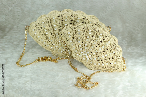 Beige crocheted shoulder bag in the shape of a seashell with a gold-colored handle sits on a white fluffy blanket. The bag features pearl bead details.