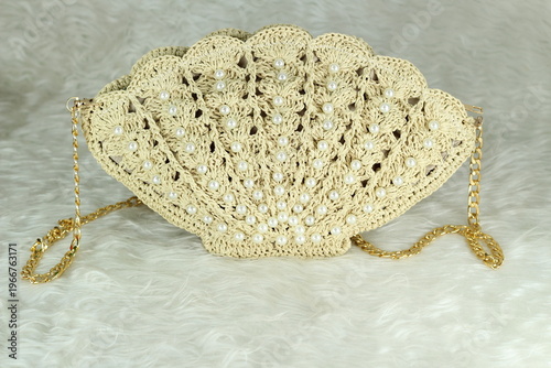Beige crocheted shoulder bag in the shape of a seashell with a gold-colored handle sits on a white fluffy blanket. The bag features pearl bead details.