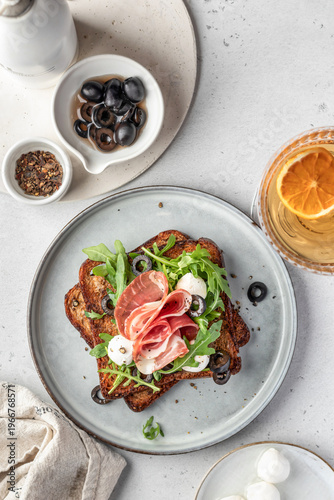 Roasted toasts with prosciutto ham, mozzarella balls, arugula and sliced olives on ceramic plate on white table background top view. Delicious breakfast