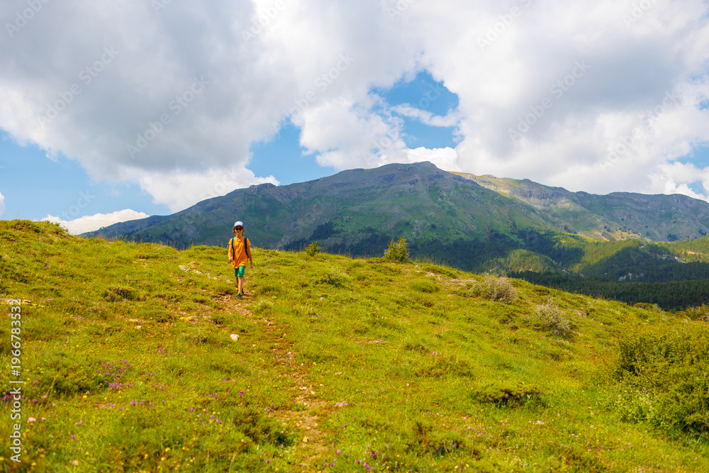 Obraz premium hiking in the mountains. A little boy with a backpack walks along a path against a background of mountains and clouds. Active healthy lifestyle on weekend hike journey.
