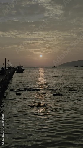 Video of a sunset view at the pier, there is a boat docked with the sunset and island in the background.