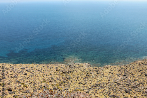 A serene and idyllic coastal view from the natural park near Almeria, Andalusia. The dark, textured volcanic soil and rocks in the foreground provide a dramatic contrast to the tranquil, deep blue sea