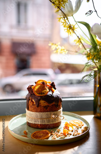 Chocolate-glazed Easter bread decorated with dried oranges and nuts on a plate by a sunlit window.