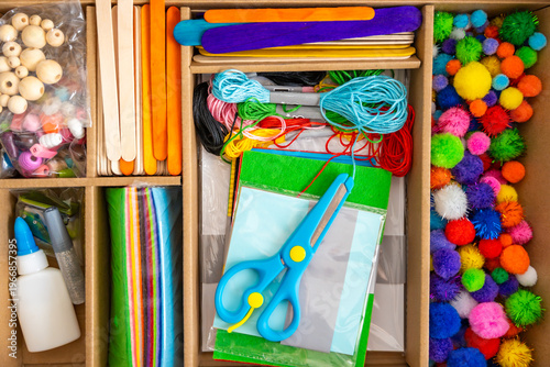 Colorful craft supplies kit with scissors paper threads beads and pom poms arranged in box on table. Creativity development fine motor skills and kids education concept.