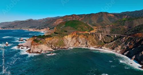 Bixby Creek Bridge and turquoise cove at Big Sur coast. Wide aerial perspective of the arched bridge with ocean waves and rocky shoreline in foreground.