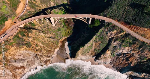 Bird's eye view of Bixby Creek Bridge with red car. Top down aerial perspective of an arched concrete bridge spanning a canyon over a beach in Big Sur, California.