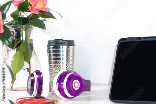 Bild auf Leinwand Top view of a workspace with black tablet, purple headphones, pink flowers and metal cup on white background