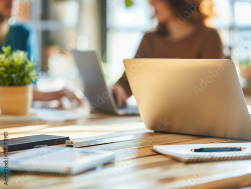 Modern Workspace with Laptops and Office Supplies,A well-lit office setting with two laptops open on a wooden table, surrounded by notebooks, a smartphone, and a potted plant