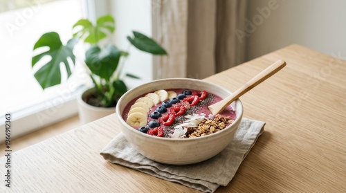 A bowl of colorful fruit and granola sits on a wooden table by a window.
