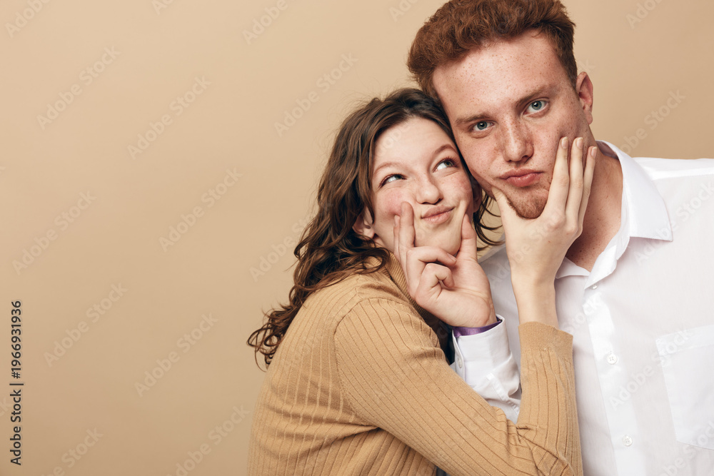 Naklejka premium Young couple with playful expressions posing together on beige background. Woman in brown sweater and man in white shirt show fun and affection. Concept of friendship and joy.