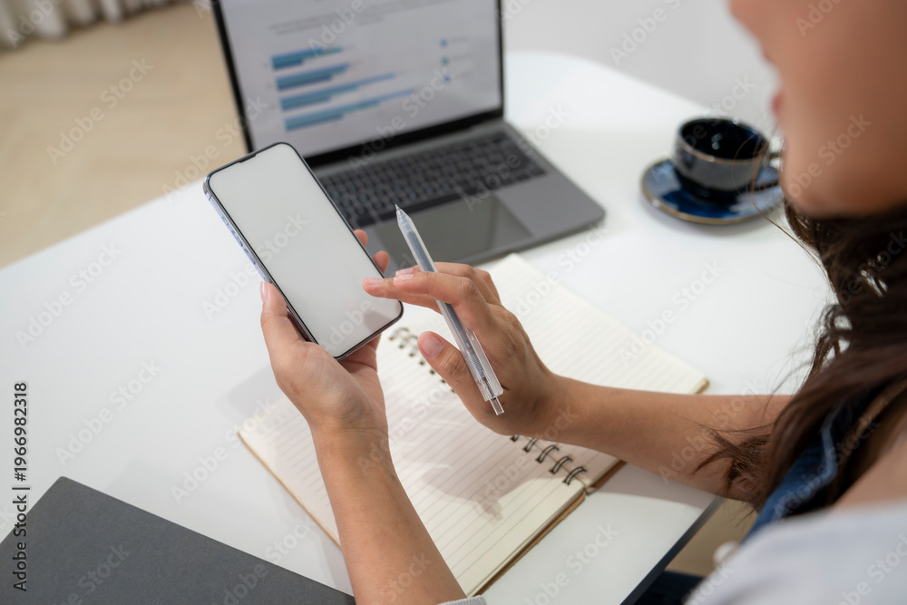 Fototapeta premium Person Using Smartphone and Writing in Notebook at Workspace