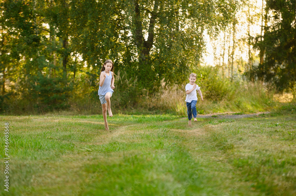 Fototapeta premium Happy Siblings Running Barefoot on Green Grass in Summer Park. Childhood Freedom and Joy Concept. Little Boy and Girl Racing on Meadow at Sunset Golden Hour. Rural Vacation, Active Lifestyle