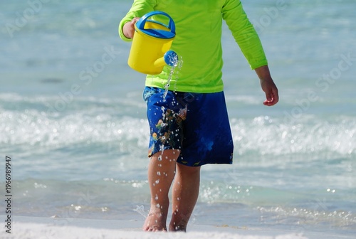 A young boy wearing bright swimwear and pouring water from a yellow watering can into the surf.