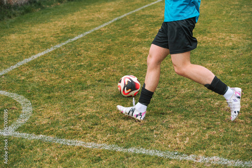 Young athlete kicks soccer ball on grass field. Grass is green with faded white lines. Player wears blue shirt and black shorts. Shoes are white with colorful accents. Game appears to be in progress