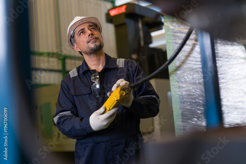 Professional male engineer wearing safety uniform operating remote control pendant for crane lifting heavy equipment in industrial factory interior workshop.