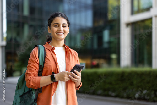 Young indian woman student wearing an orange shirt and backpack, smiling at camera while checking her smartphone on a modern university campus setting