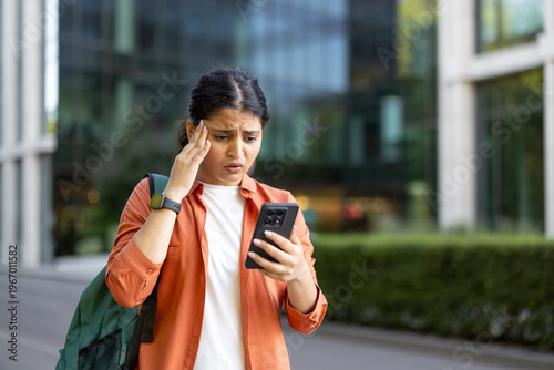 Young woman standing outdoors in a city, experiencing stress and a headache after seeing bad news on her mobile phone, feeling worried, frustrated, and shocked