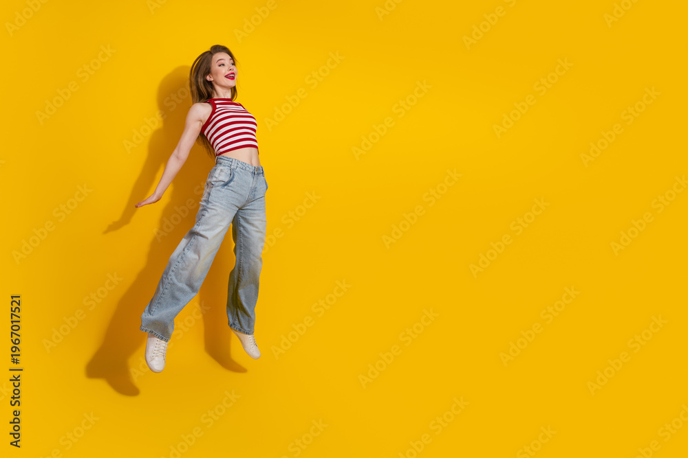 Fototapeta premium Cheerful young woman in striped top posing playfully on a vibrant yellow background, showcasing casual and trendy summer style
