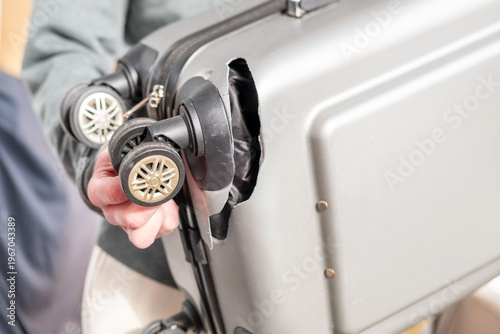 Passenger holding damaged silver suitcase with extensive broken wheel, corner, cracked hardshell luggage, exposed wheel mechanism, travel inconvenience, baggage damage, travel mishap, close-up view