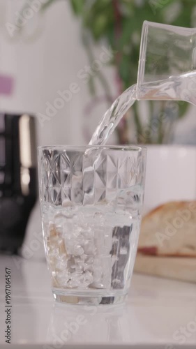 Vertical shot of pouring clean still water into a minimalist glass. Preparation for a daily health routine in a bright, modern kitchen interior.