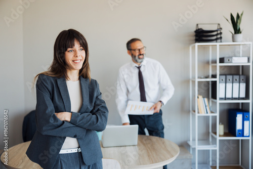 Young professional businesswoman smiling at the camera with an older male colleague working at his desk in the background.
