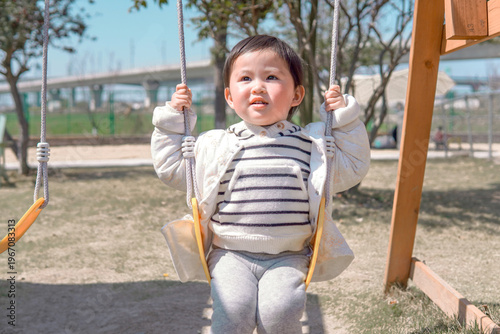 Happy Asian toddler girl enjoying a swing at an outdoor playground. Cheerful child having fun and laughing in the sunny park.