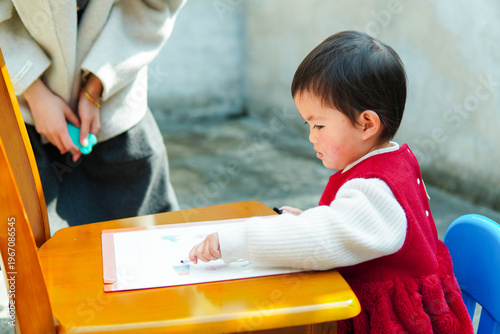 Cute Asian toddler girl drawing with a crayon at a small desk. Little child focused on creative artwork and early art education at home.