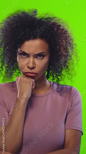 Young African American woman with afro hairstyle in casual clothes expresses doubt and deep thought with hand on chin against green backdrop. Emotion, pondering, reflection idea.