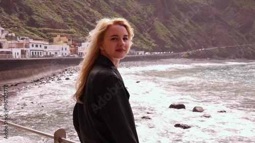 Side profile of young blonde woman enjoying fresh air by Atlantic ocean with scenic coastal village and green mountain cliffs in Tenerife. Ideal for travel and lifestyle projects