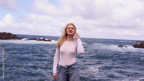 Beautiful young blonde woman standing on Atlantic rocky coastline with ocean waves and cloudy sky in Tenerife, Canary Islands. Perfect for travel, lifestyle and fashion projects