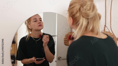 Young blonde woman applying makeup with brush while looking in round mirror at home. Perfect for beauty, lifestyle, cosmetics, morning routine and self-care themed projects